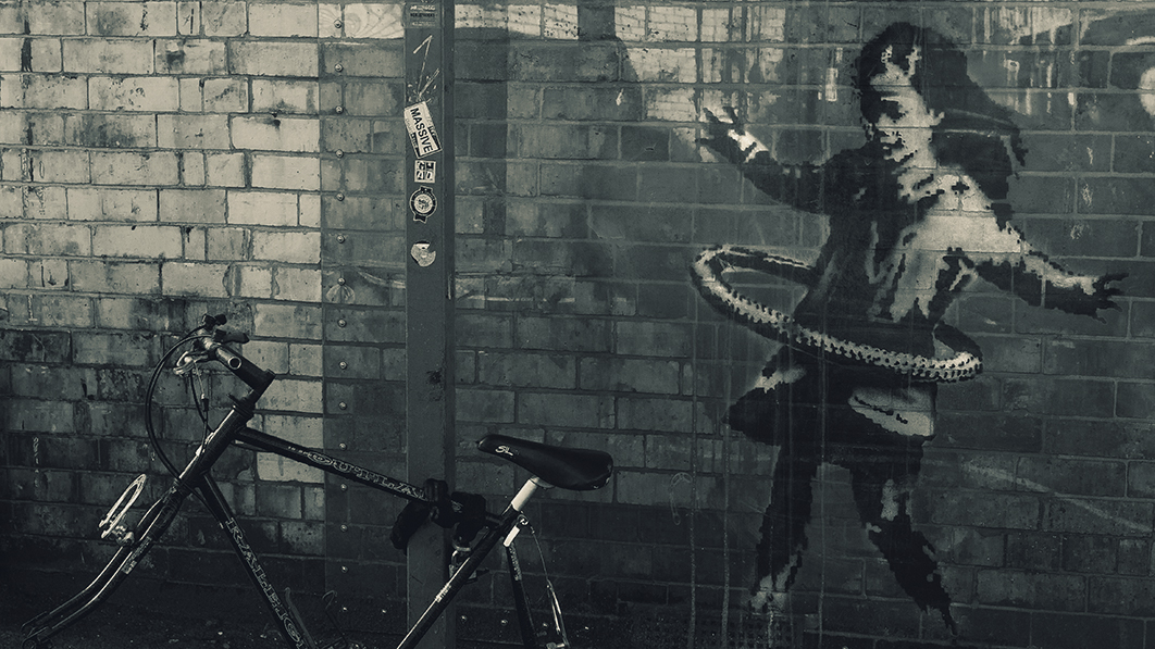 Foto einer grauen Ziegelwand mit angelehntem Fahrrad, auf der Wand: mutmaßlich ein Banksy-Bild: Mädchen mit Hula-Hoop-Reifen