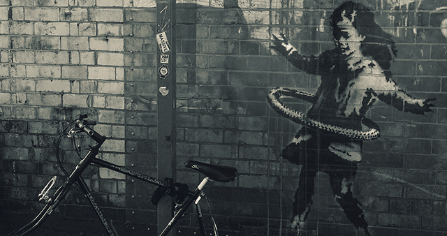 Foto einer grauen Ziegelwand mit angelehntem Fahrrad, auf der Wand: mutmaßlich ein Banksy-Bild: Mädchen mit Hula-Hoop-Reifen
